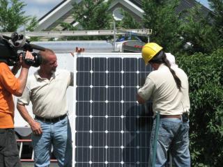 A worker demonstrates solar installation while a camera crew looks on