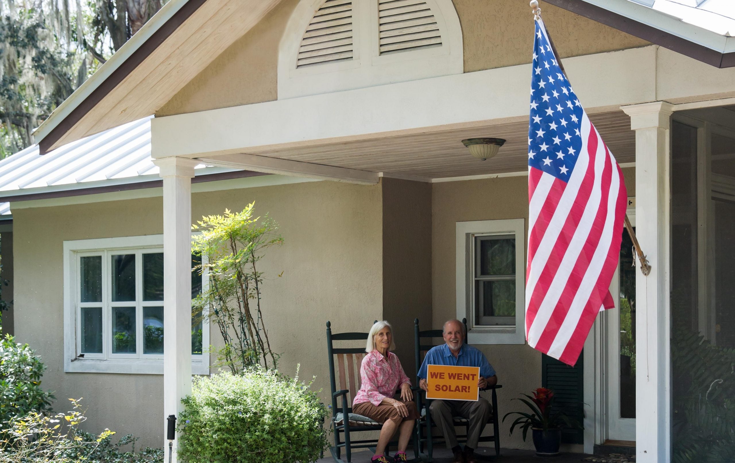 A couple sits on their porch with a sign that says "WE WENT SOLAR" in front of an American flag
