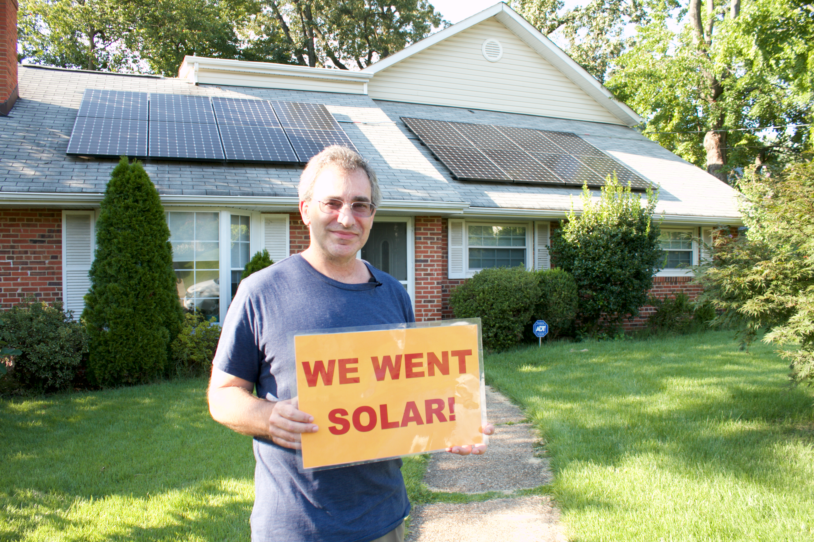 Virginia solar homeowner in front of their home with solar panels. Homeowner has a yellow sign with red text saying we went solar.