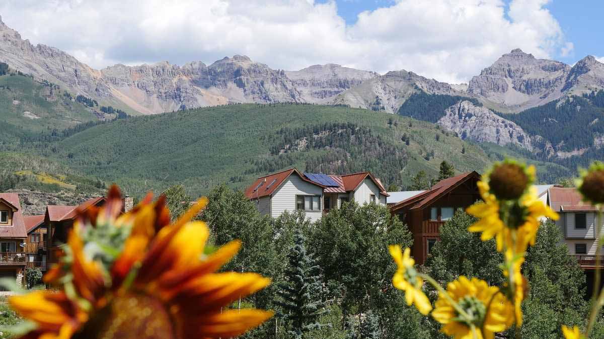 A house with roof top solar panels. There are sunflowers in the forefront and mountains in the background.