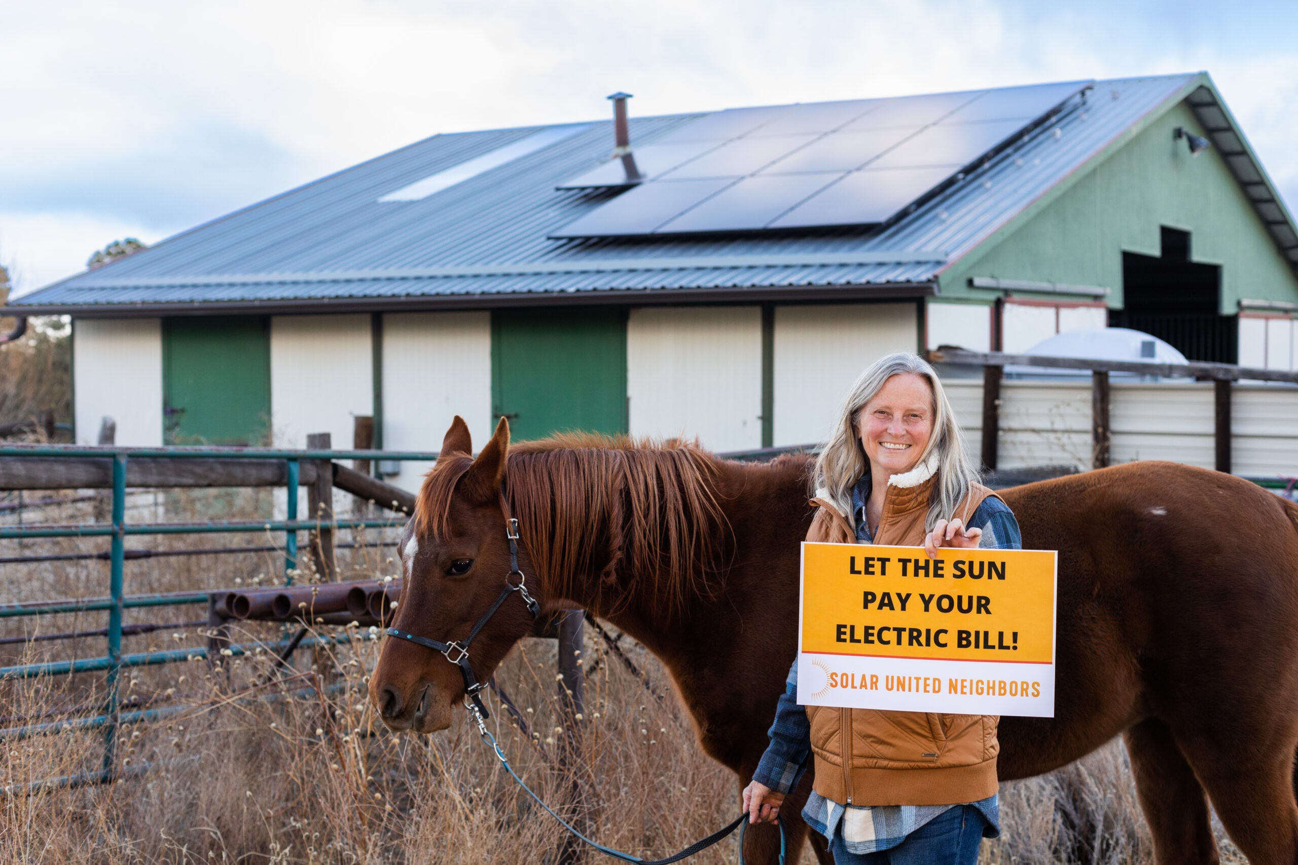 Co-op member holding "Let the sun pay yoour electicity bill!" sign with a brown horse next to them. Solar array on barn in the background.