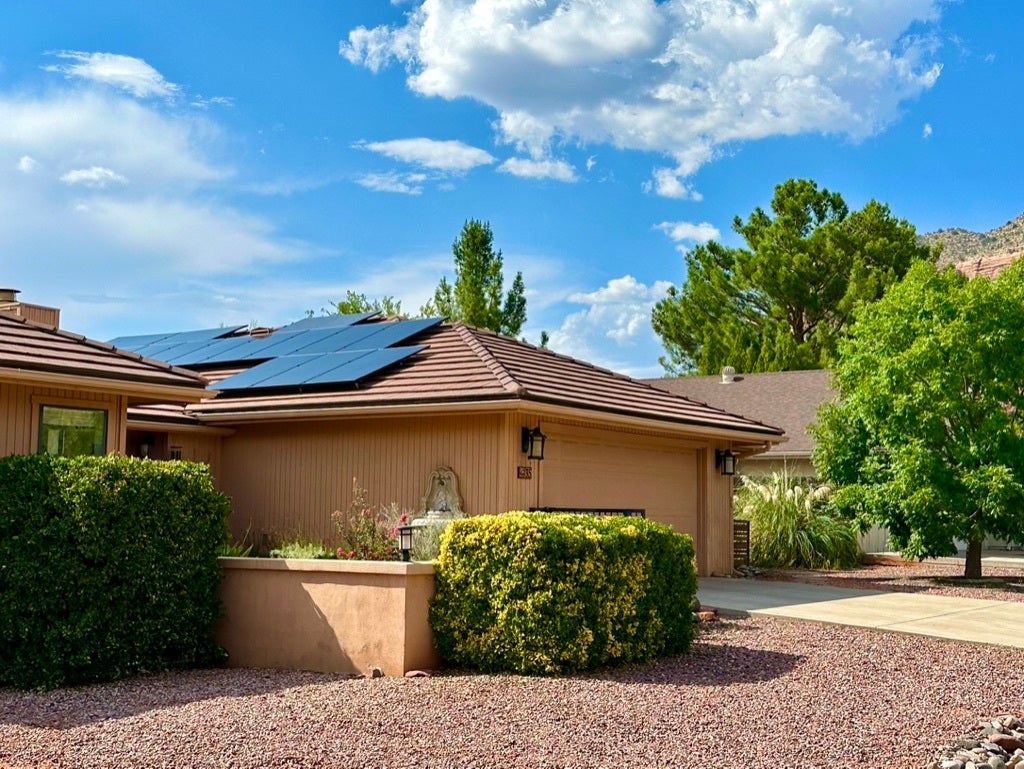 Photo of a brown house with rooftop solar panels. Clear blue sky in the background and green foliage around the house.