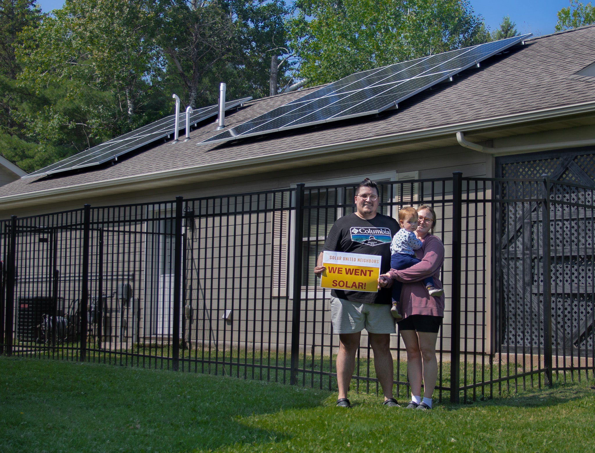A family with a small child hold a "we went solar!" sign in front of their house with rooftop solar panels.